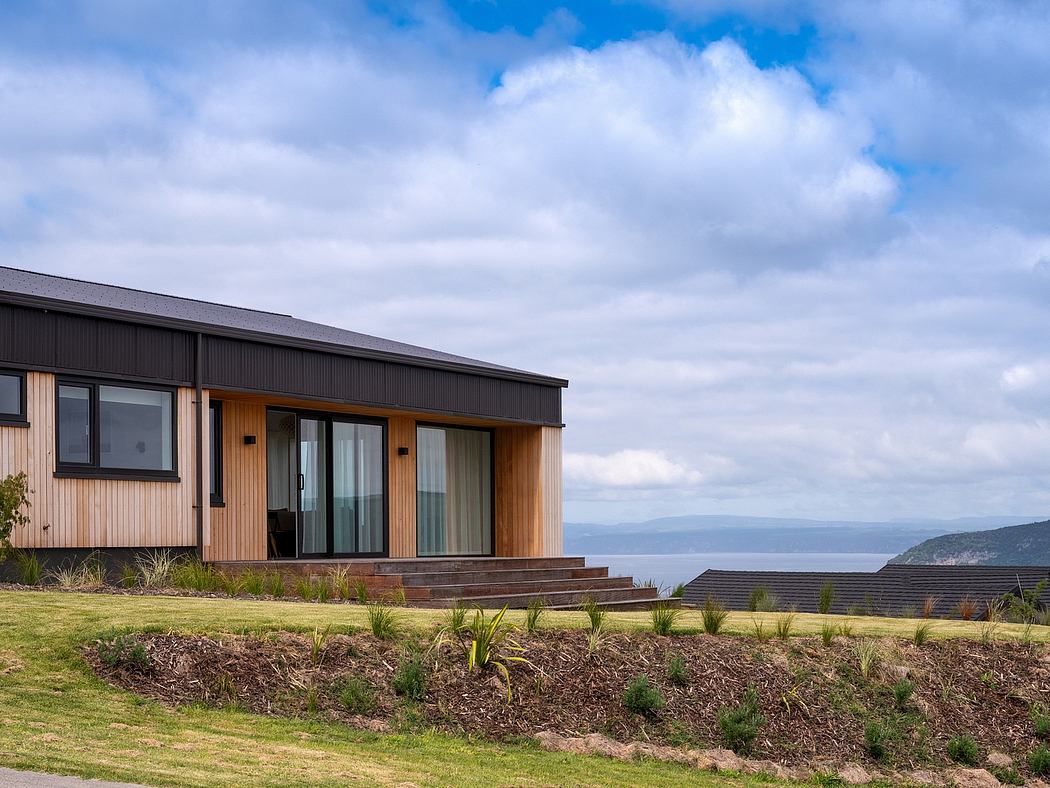 A modern wooden house with large windows overlooking a grassy landscape and distant hills.