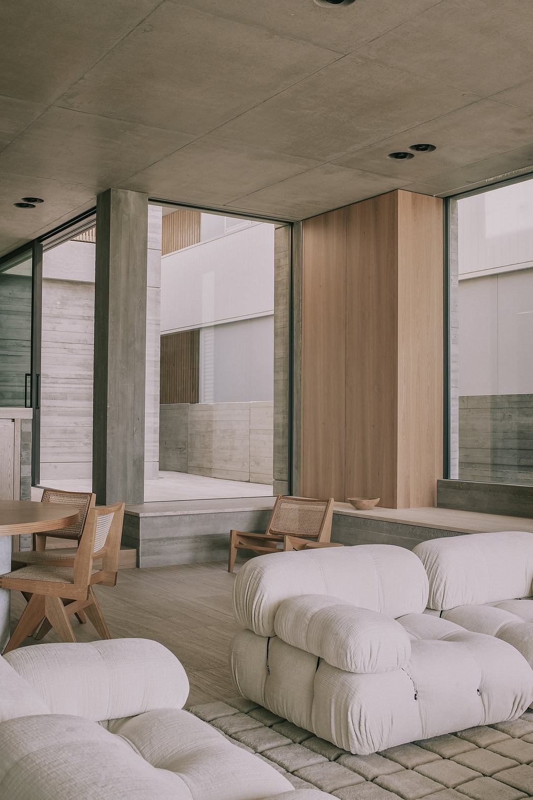 Minimalist concrete and wood interior with large windows, cushioned seating, and geometric patterns.