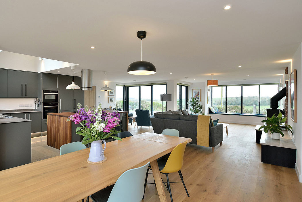 Modern, open-plan kitchen and living area with wooden dining table, pastel chairs, and floor-to-ceiling windows.