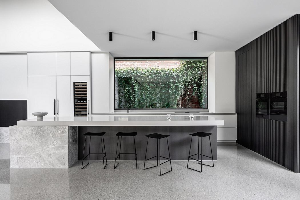 Minimalist kitchen with sleek marble countertop, black stools, and large window overlooking greenery.