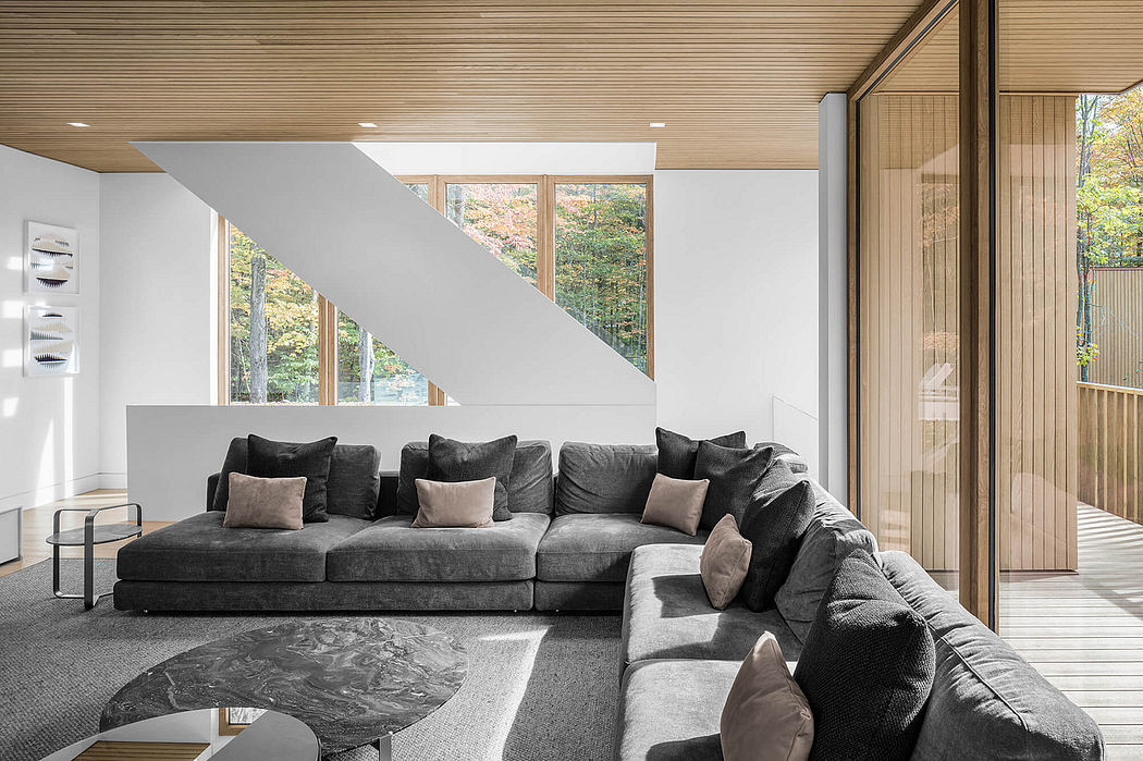 Spacious living room with large windows overlooking a lush, wooded landscape. Minimalist design with modern gray couch and decor.