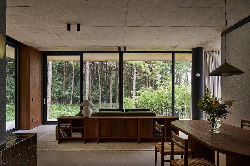 Open-plan living space with wood-paneled walls, large windows overlooking a forested landscape, and a dining area with a rustic wooden table and chairs.