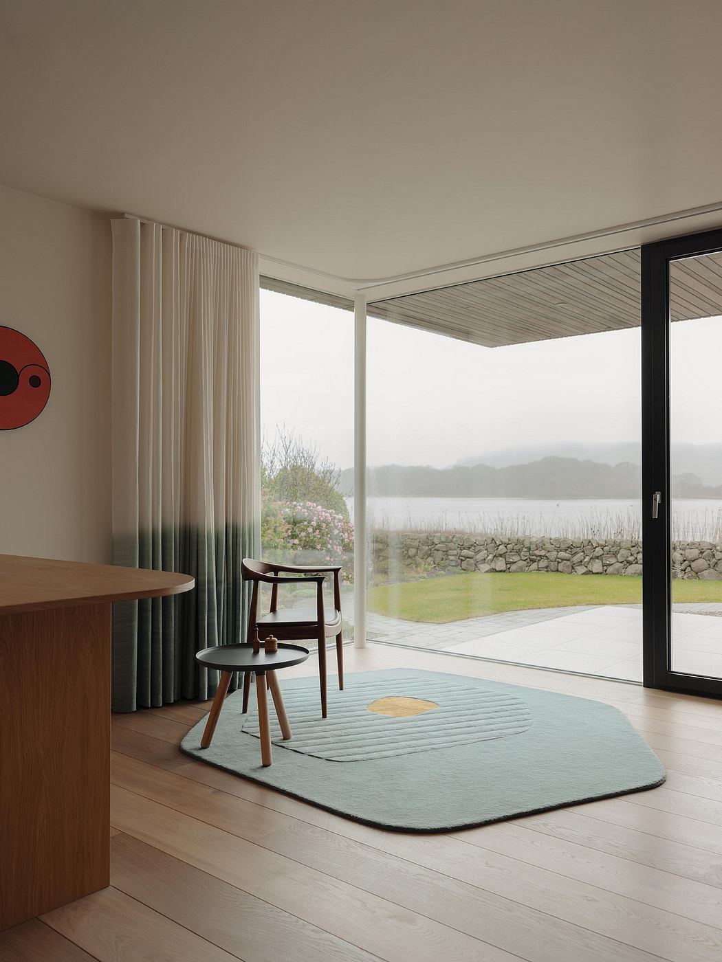 Minimalist living room with wooden furniture, gray rug, and large window overlooking a lake.