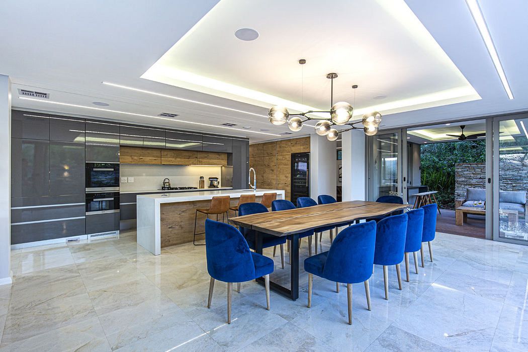 Spacious dining area with sleek kitchen, wooden table, blue velvet chairs, and pendant lighting.