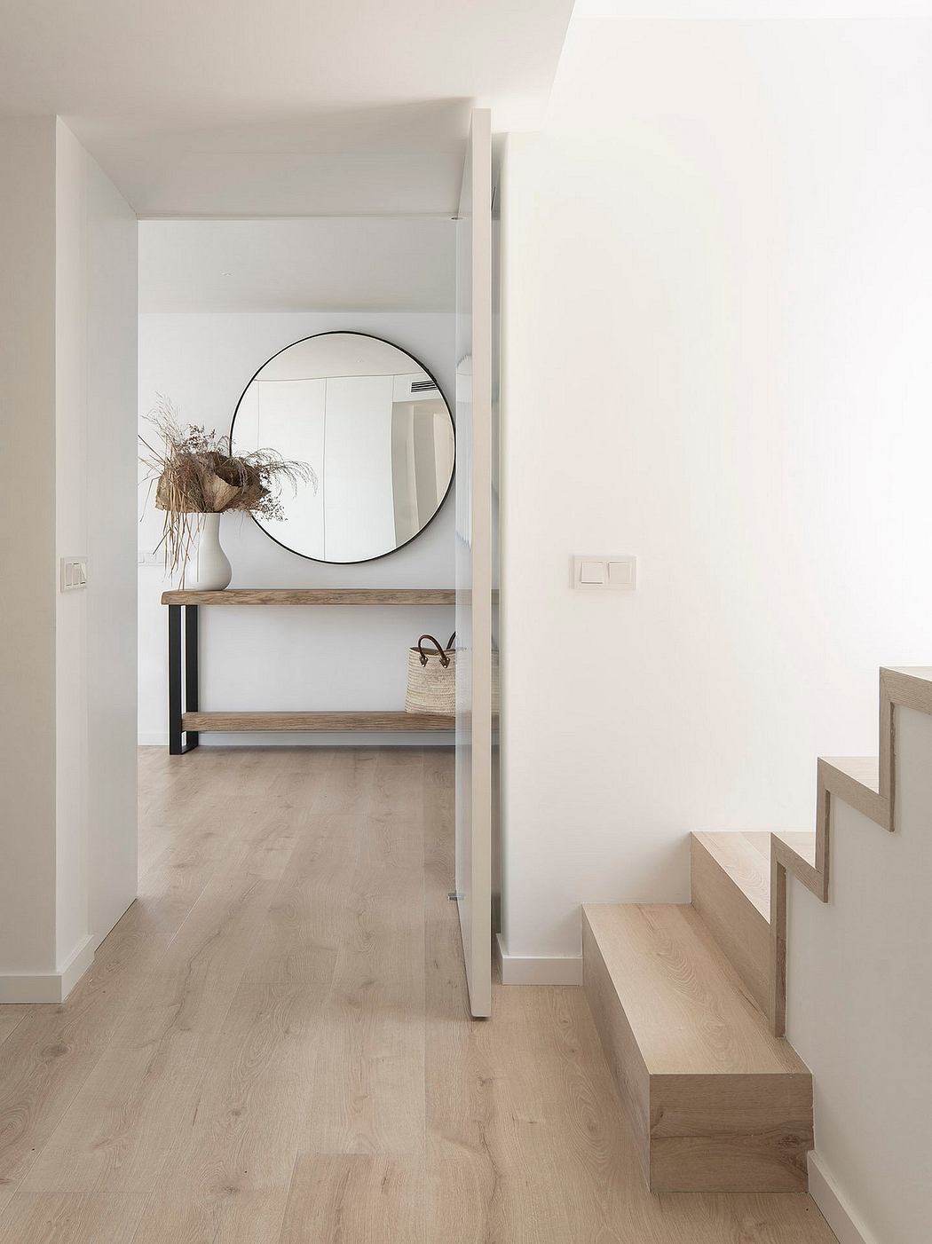 Minimalist entrance with wooden stairs, shelving unit, and round wall mirror.