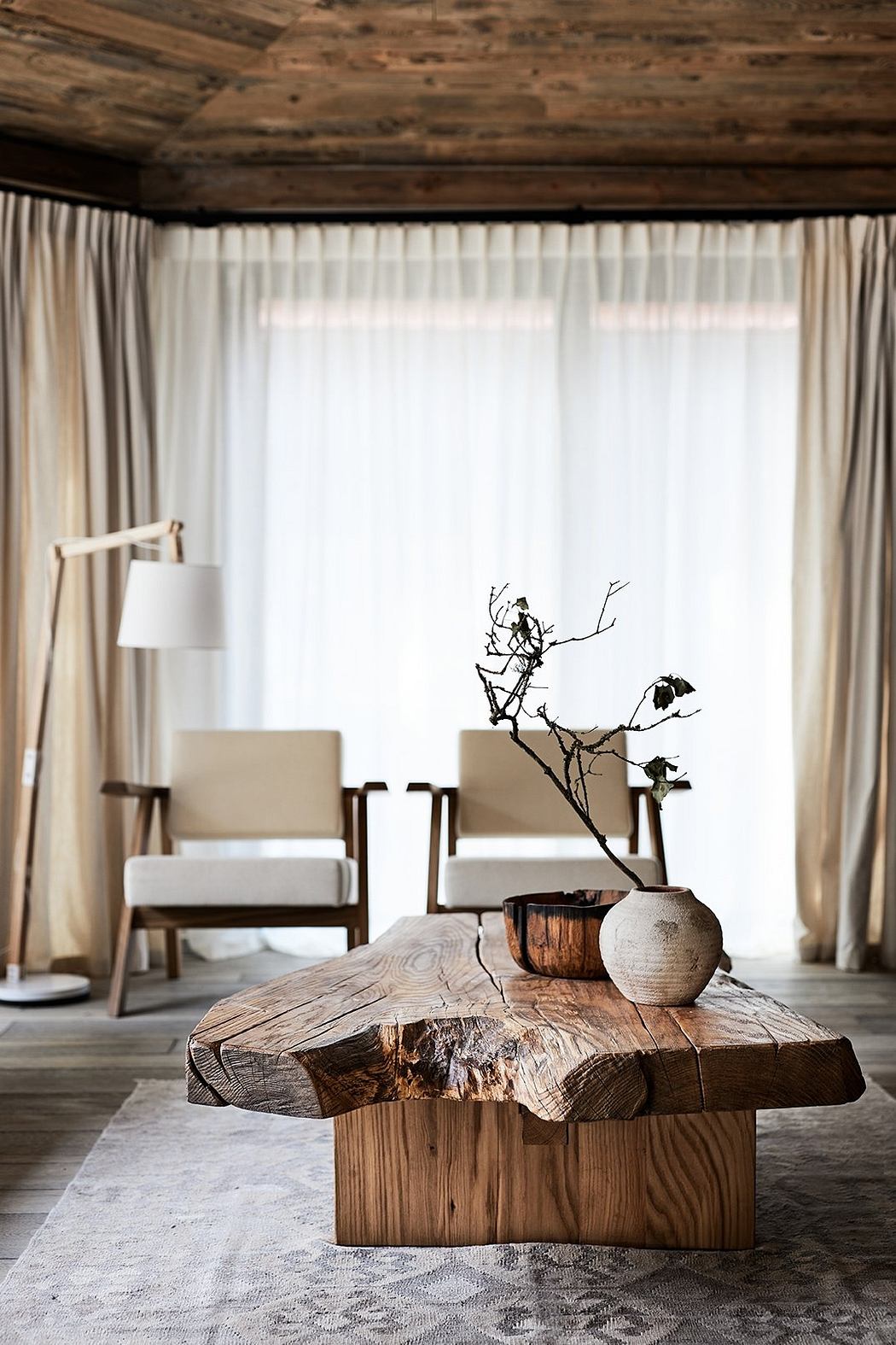 A rustic living room with wooden ceiling beams, white curtains, and a handcrafted wooden coffee table.