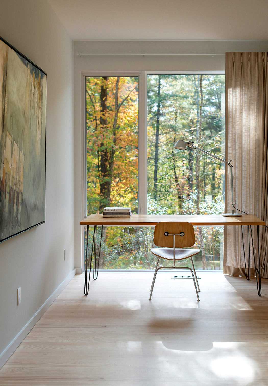 A cozy home office with a wooden desk, mid-century chair, and large window showcasing autumn foliage.