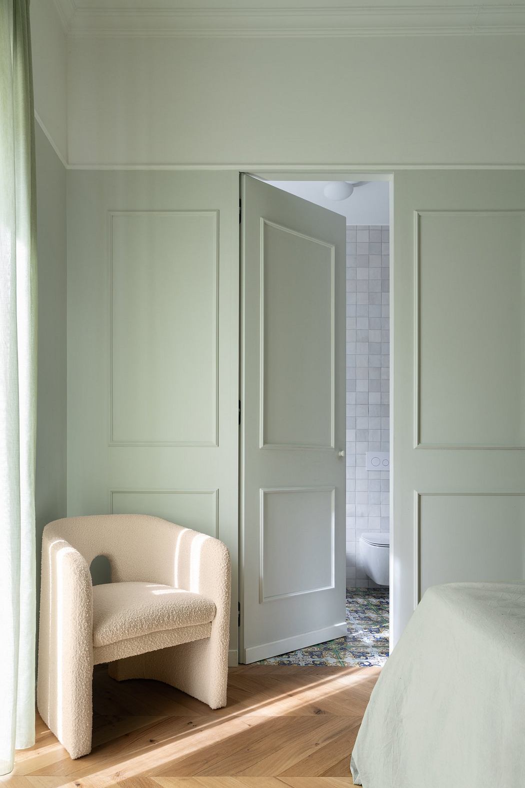 Cozy, modern bedroom with plush armchair, paneled walls, and glimpse of tiled bathroom.