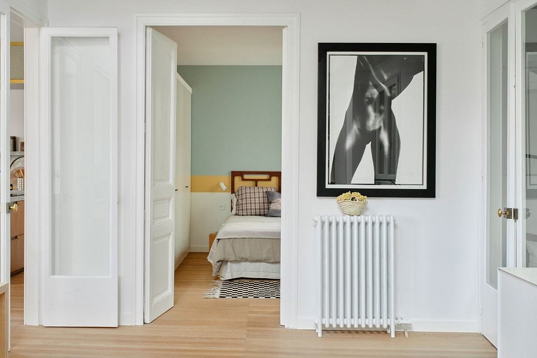 Minimalist bedroom with white walls, wooden floors, and a black-framed nude artwork.