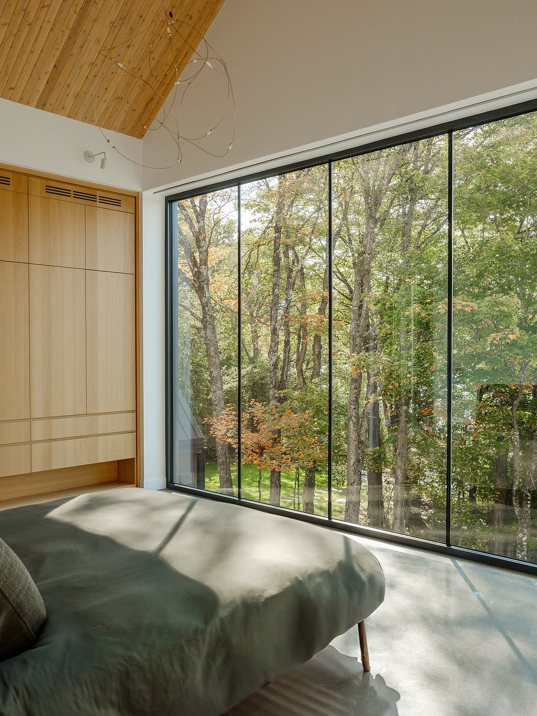 A cozy bedroom with a serene forest view, featuring floor-to-ceiling windows and a wooden ceiling.
