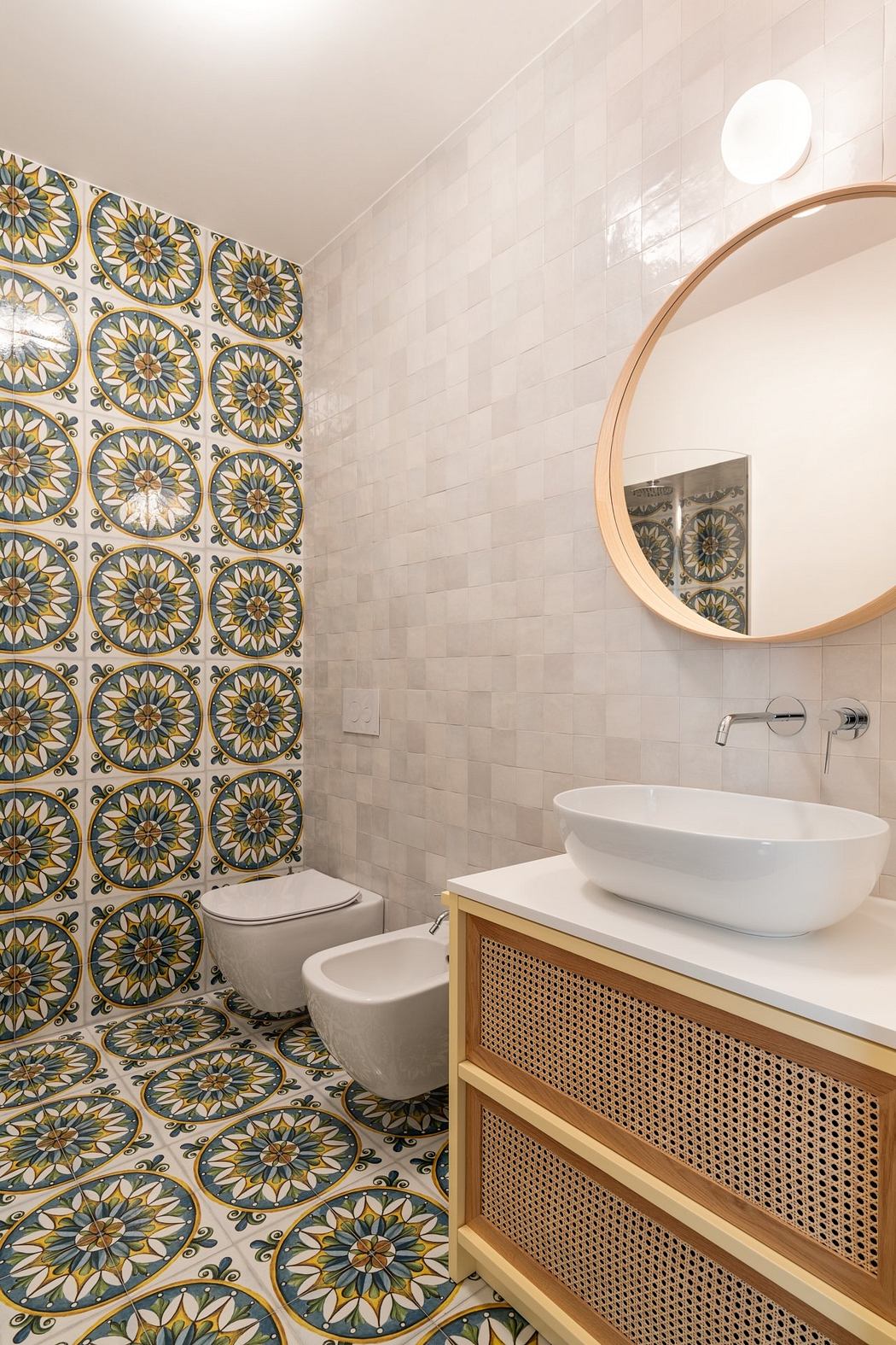 Ornate tile pattern on walls, wooden vanity with rattan drawers, round mirror in bathroom.