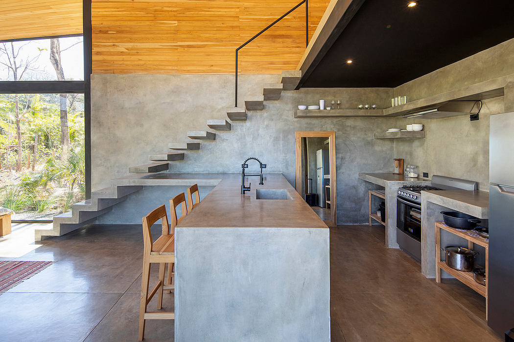 A modern, open-concept kitchen and living room with concrete walls, wooden ceiling, and floating stairs.