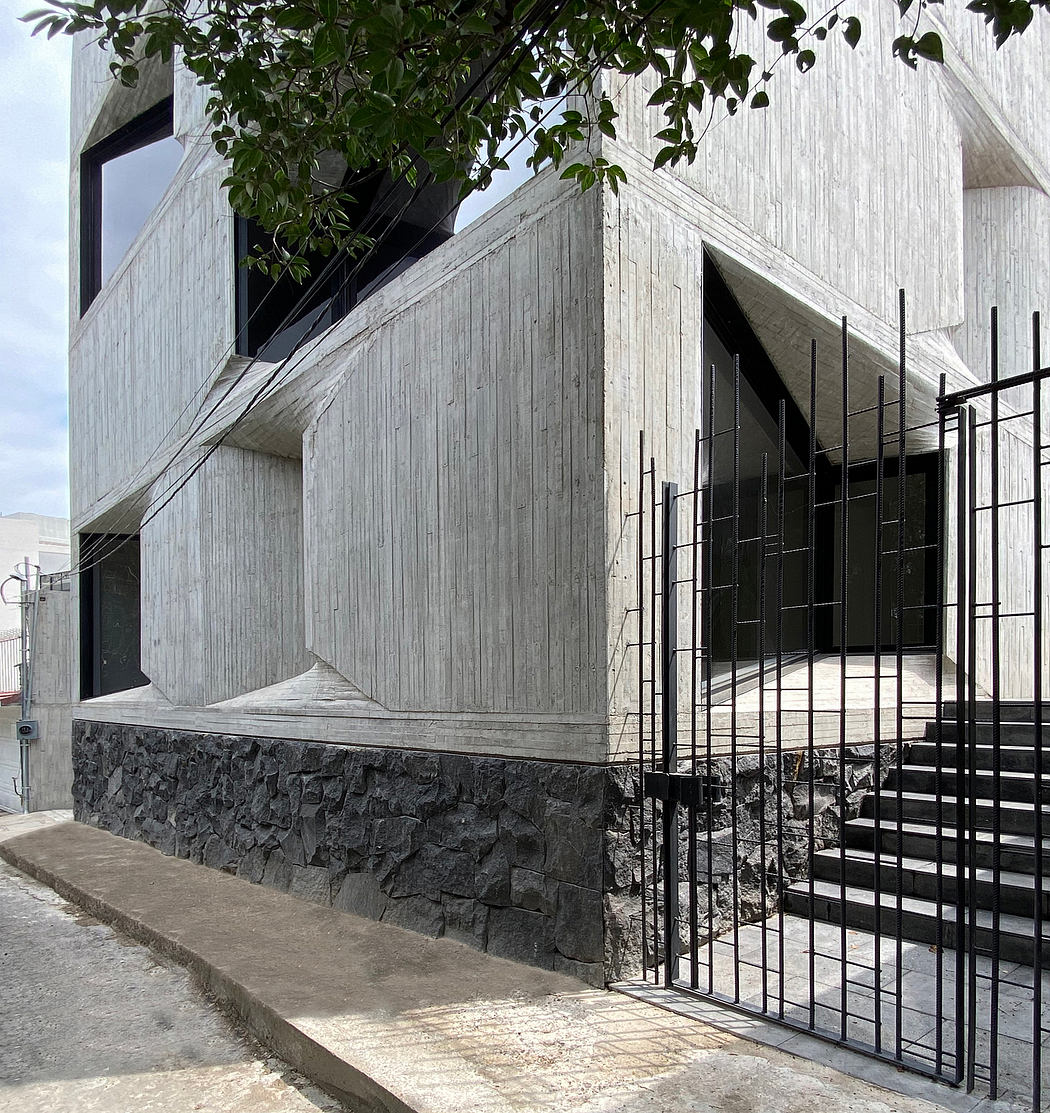 A modern, angular concrete building with a stone base and metal railings, framed by lush greenery.