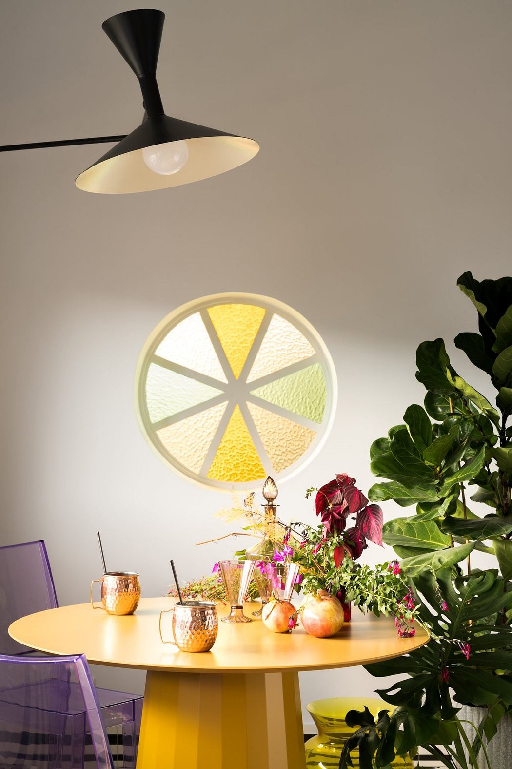 A modern dining area with a circular stained glass window, lush potted plants, and a round yellow table.