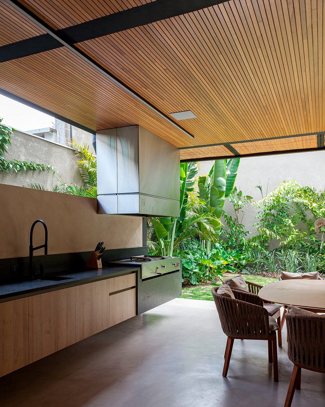 Sleek outdoor kitchen with wooden slat ceiling, lush tropical foliage, and modern furniture.
