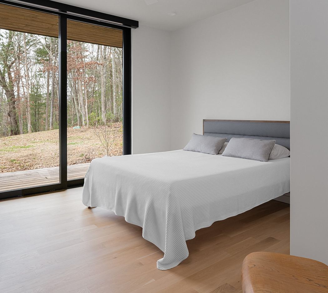 A minimalist bedroom with a large window overlooking a wooded landscape, featuring a gray upholstered headboard and hardwood floors.