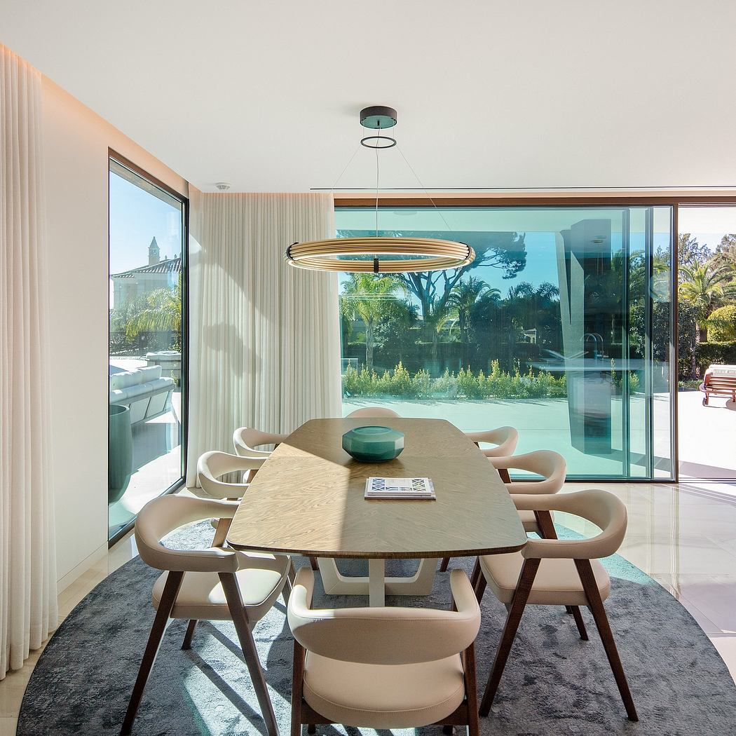 A modern dining room with wooden table, chairs, and pendant lamp; large windows overlooking lush greenery.
