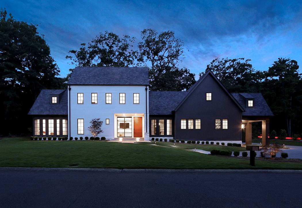 Elegant two-story house with dark exterior and well-illuminated windows, surrounded by lush trees.