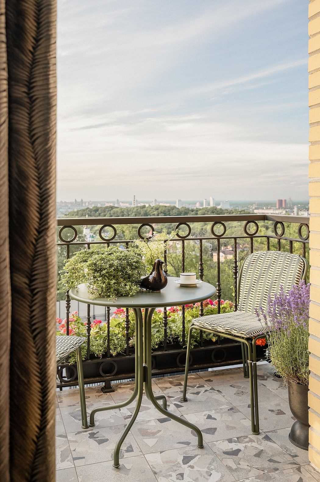 Small balcony with ornate metal furnishings, potted plants, and a view of a city skyline.