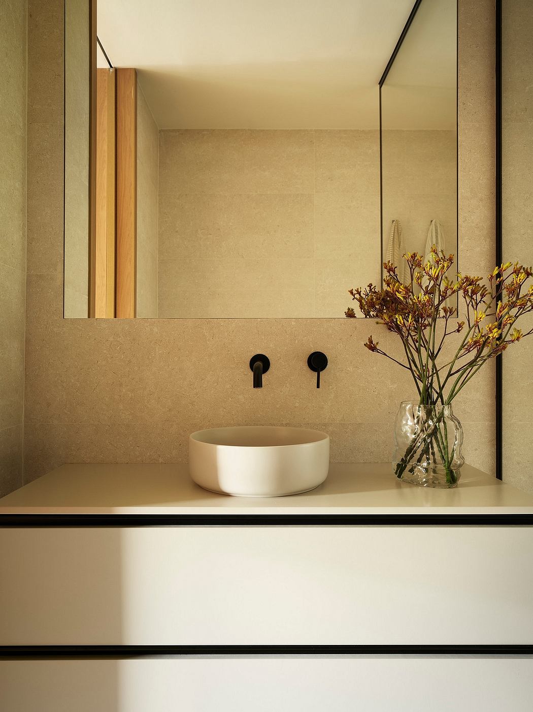 A minimalist bathroom with a white ceramic vessel sink, wooden accents, and a vase of flowers.