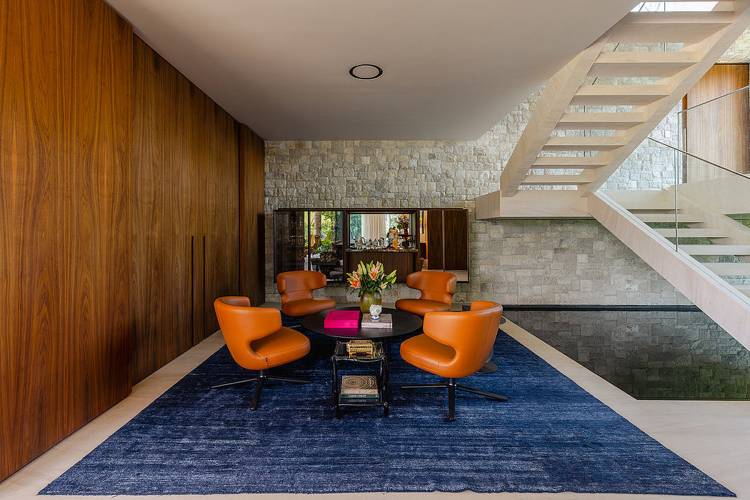 A modern living room with stone walls, wooden cabinetry, and vibrant orange chairs on a navy rug.