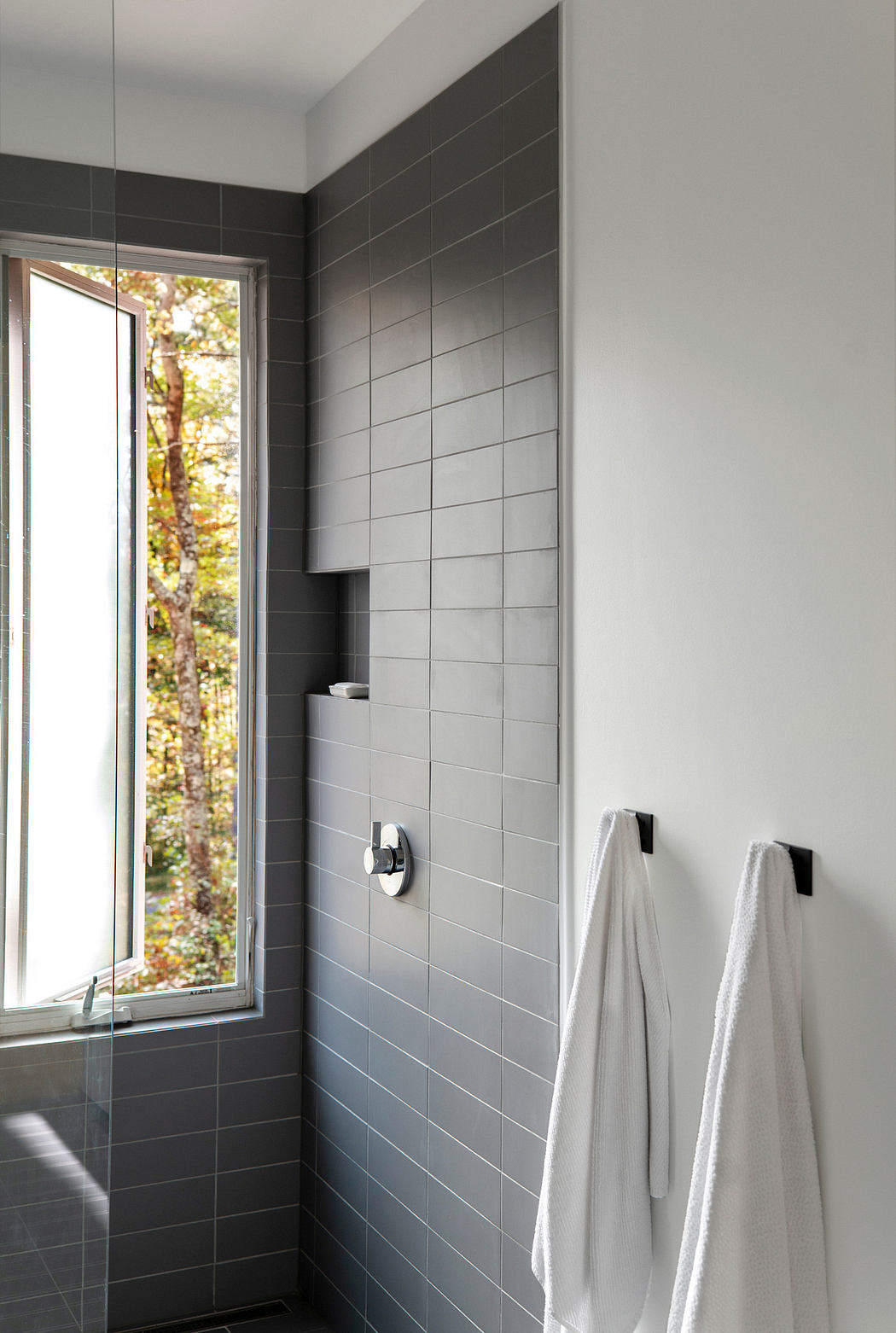 Sleek modern bathroom with gray tile walls, towel hooks, and a large window overlooking nature.