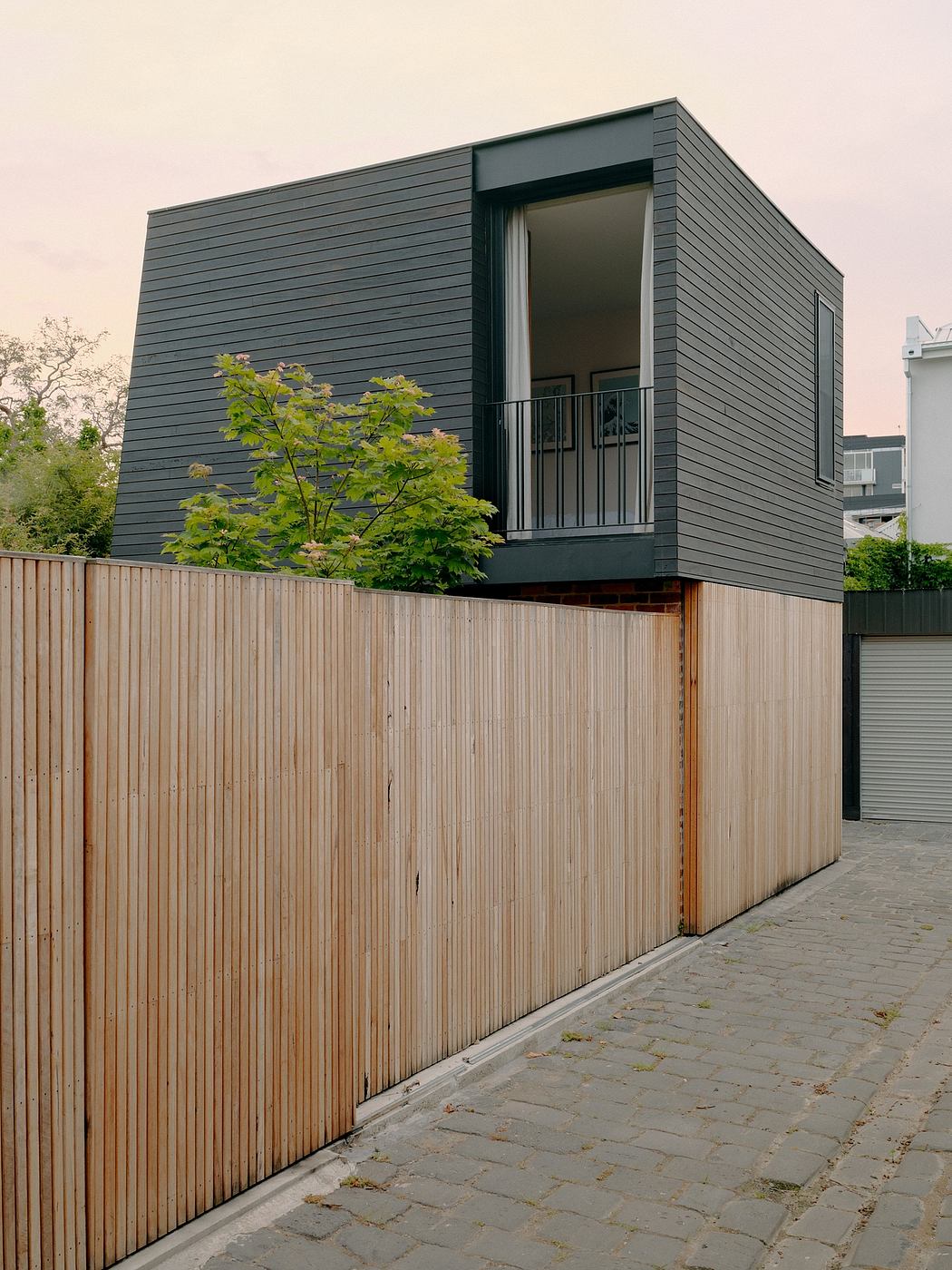 Modern, minimalist house with dark gray siding and wooden fence; balcony overlooking landscaping.