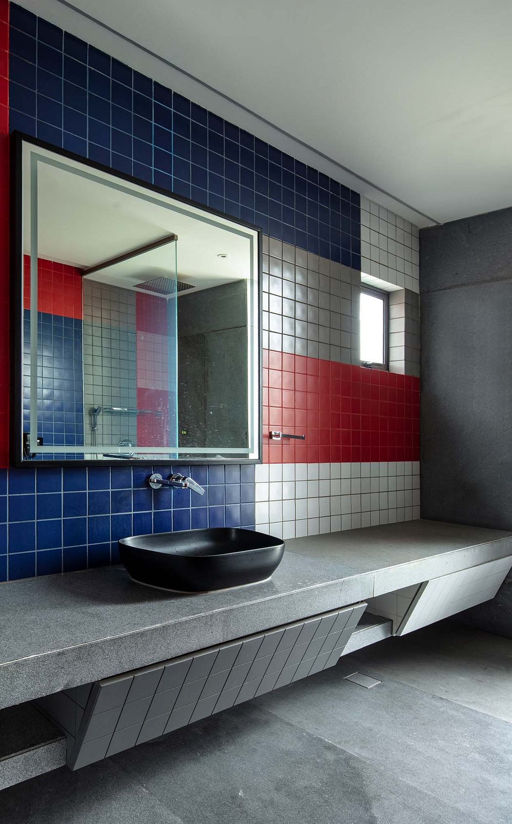 A modern bathroom with bold red and blue tiled walls, a large mirror, and a black vessel sink.