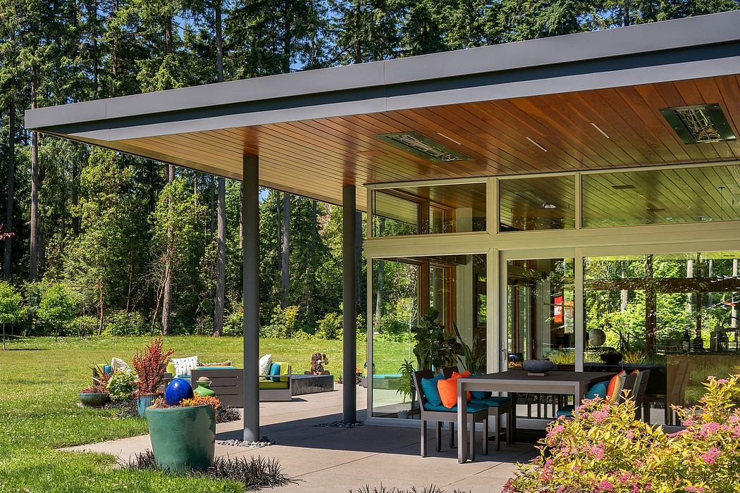 Modern wood-and-glass house with roofed patio, lush landscaping, and colorful furnishings.