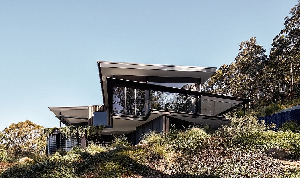 Modern glass and steel residential home perched on a sloped hillside surrounded by lush vegetation.