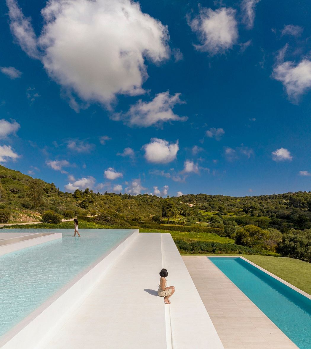 Two people enjoying the pool on a white concrete platform overlooking a lush, green landscape.