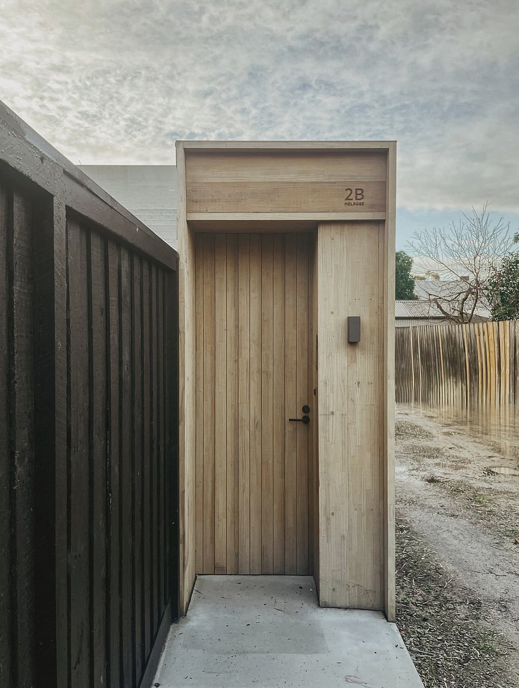 A rustic wooden building with a number 2B above the entrance and a concrete path leading to it.