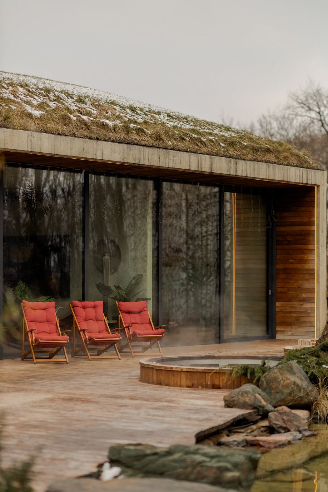 A modern cabin with a green roof and wooden patio overlooking a natural landscape. Three red cushioned chairs are arranged on the deck.