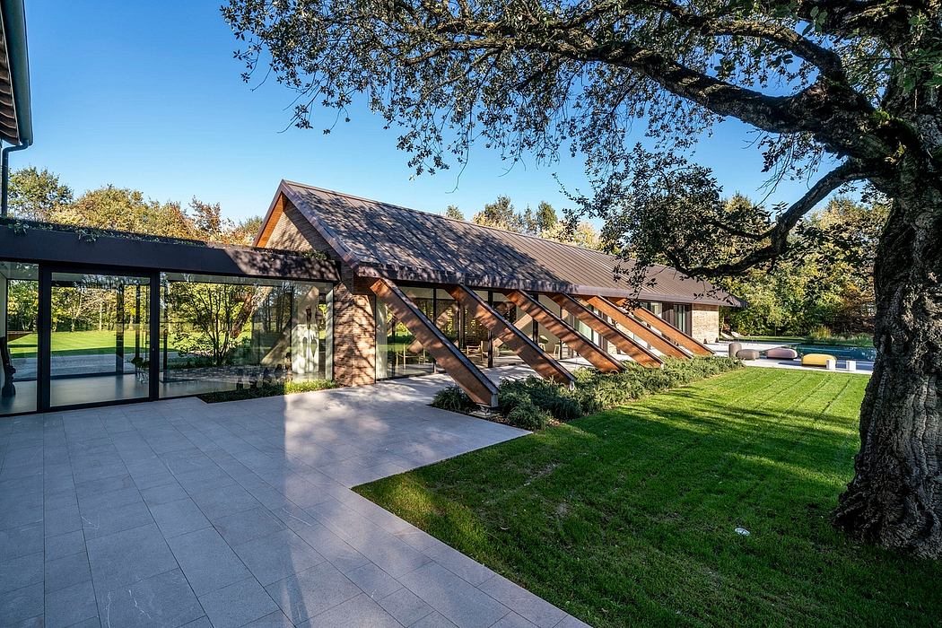 The image shows a modern, glass-encased wooden structure with a slanted roof, surrounded by lush greenery and a well-manicured lawn.