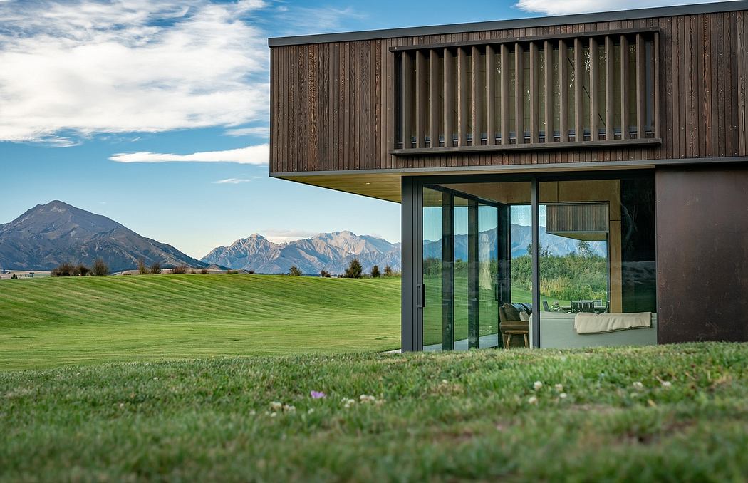 Modern wooden structure with large glass windows overlooking a grassy field and snow-capped mountains.