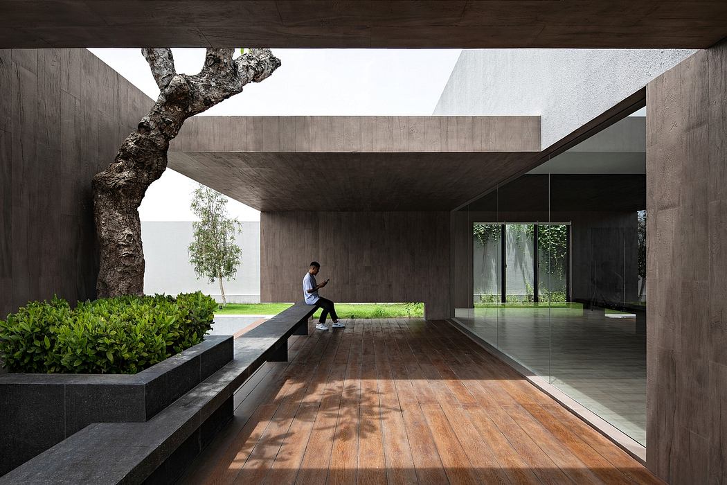A modern, minimalist outdoor pavilion with a concrete structure, wooden flooring, and lush greenery.