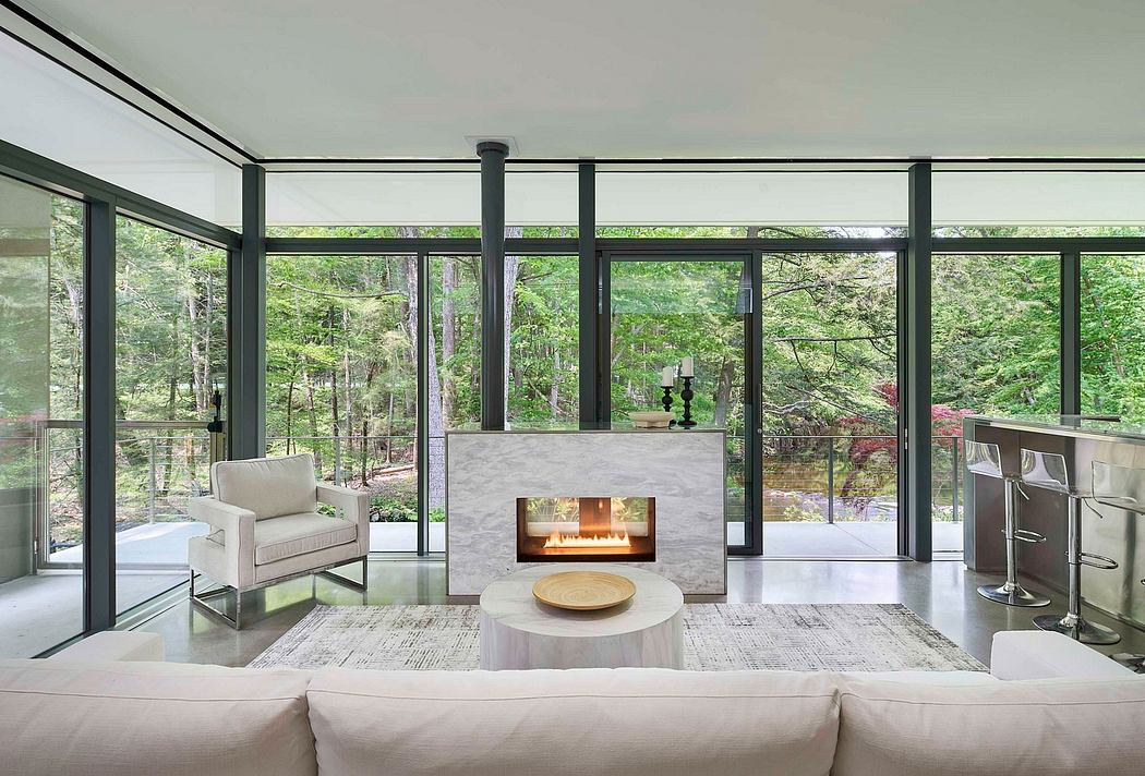 A modern glass-enclosed living room with a fireplace, cozy furniture, and lush forest views.