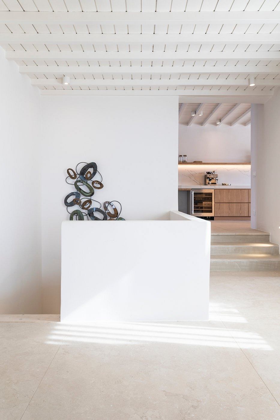 The image shows a spacious, minimalist interior with a wood-paneled kitchenette and a sculptural artwork on the wall.