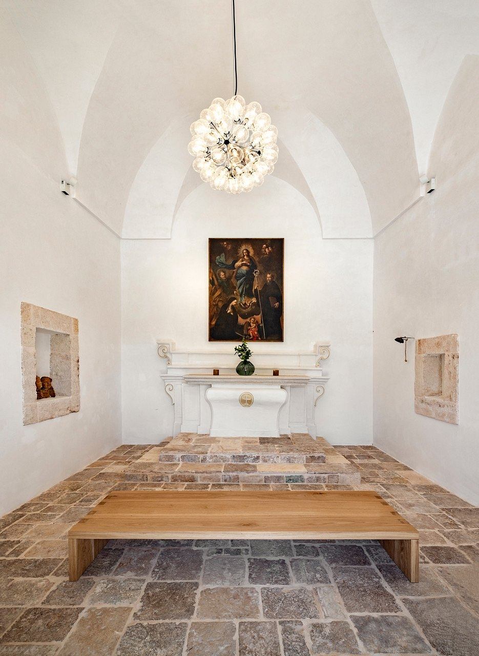 A grand, vaulted room with an ornate chandelier, religious artwork, and a rustic wooden table.