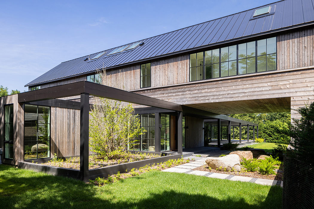 A modern, wooden structure with large windows and a covered entrance leading to a landscaped garden.