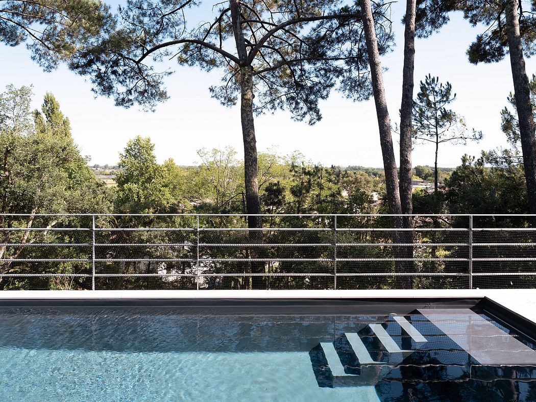 The image shows a swimming pool surrounded by a modern, minimalist architectural design with sleek metal railings and a serene forested backdrop.