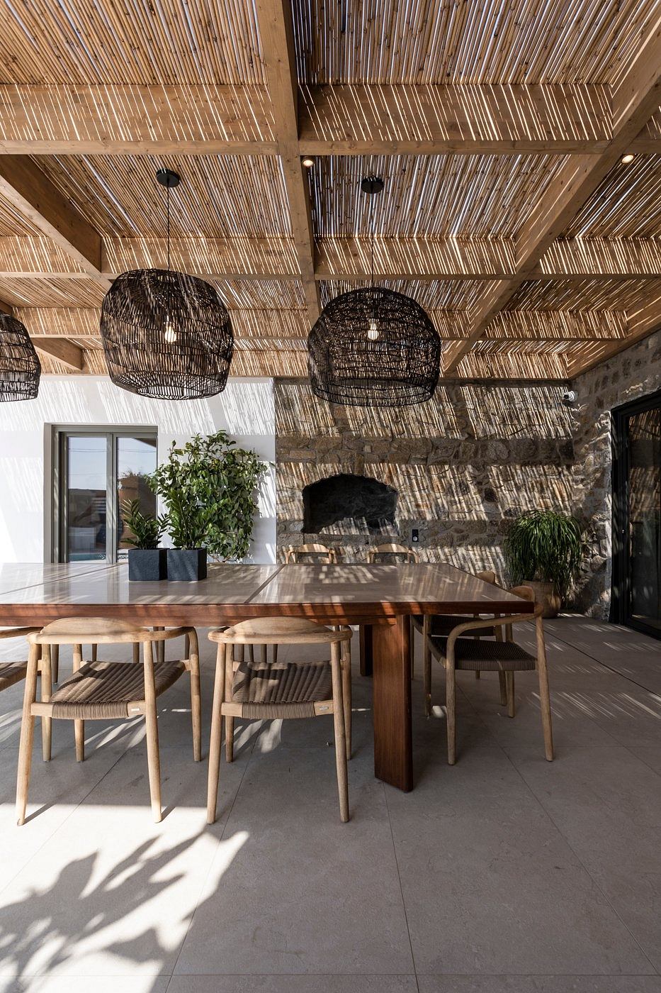 Rustic dining room with woven bamboo ceiling, wooden table and chairs, and potted plants.