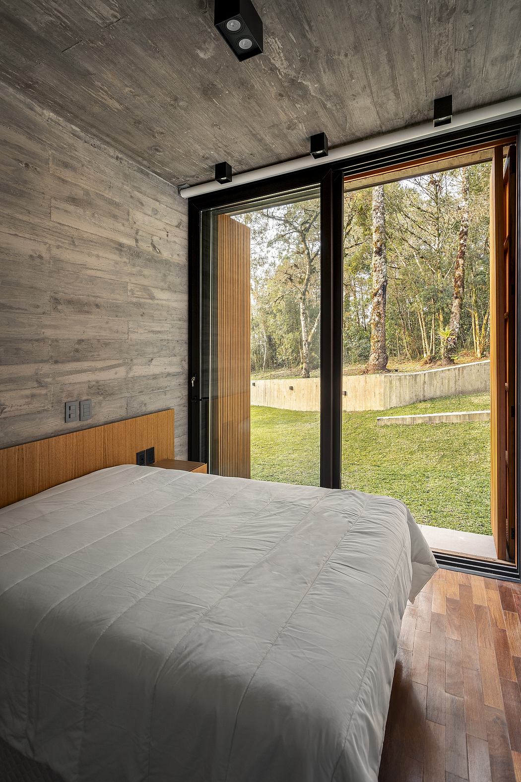 Spacious bedroom with floor-to-ceiling windows, exposed concrete walls, and wooden flooring.