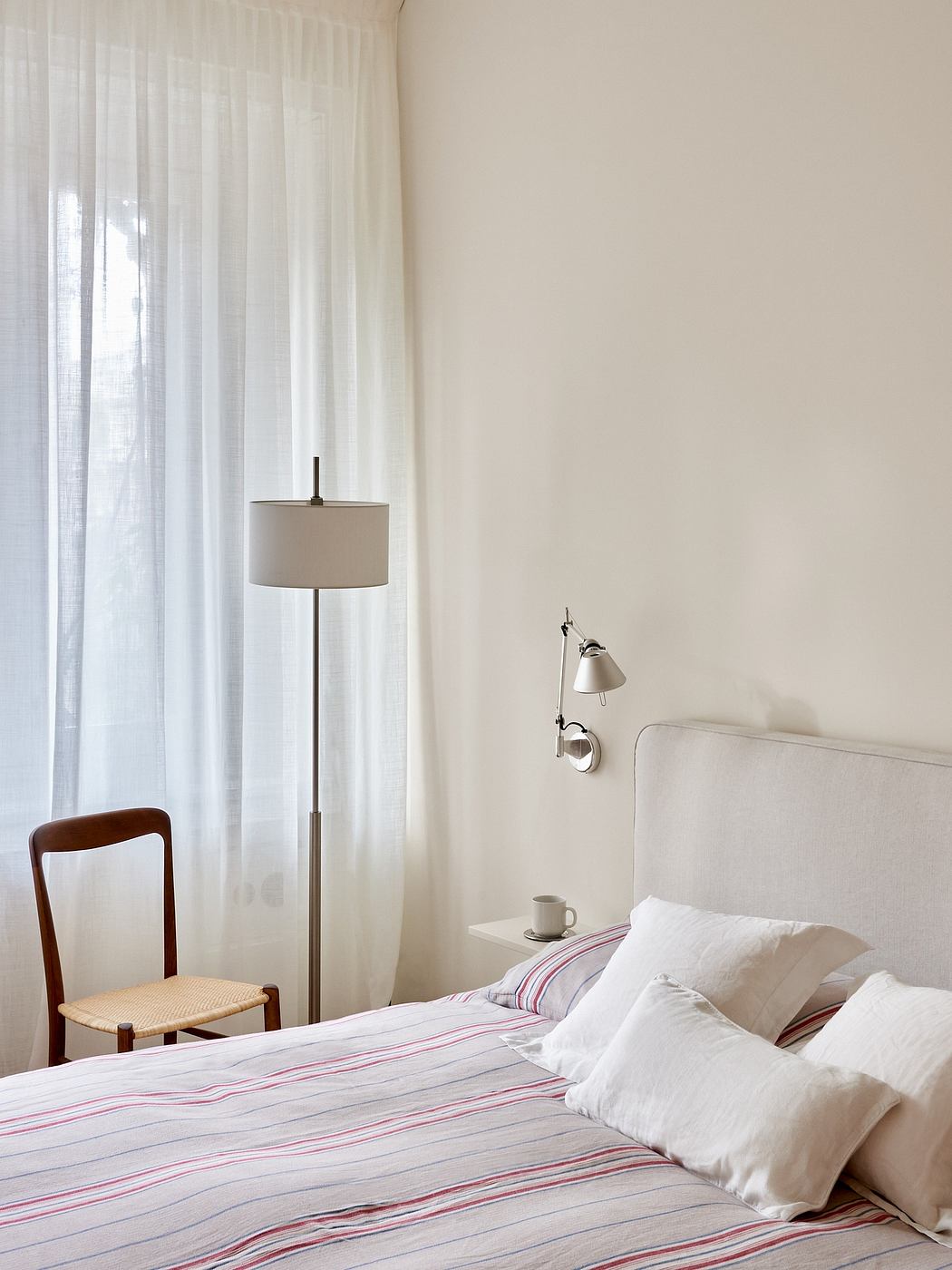 Cozy bedroom with minimal furnishings, striped bedding, floor lamp, and wooden chair.