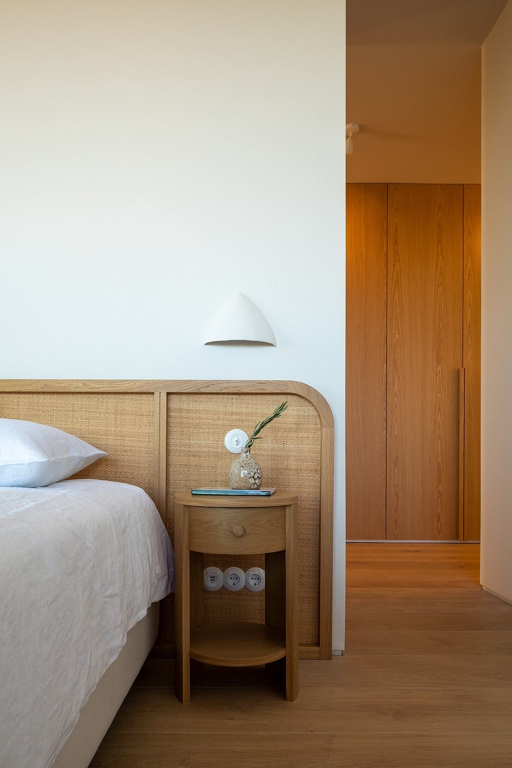 A cozy bedroom with warm wooden furniture, a bedside table, and a white wall light.