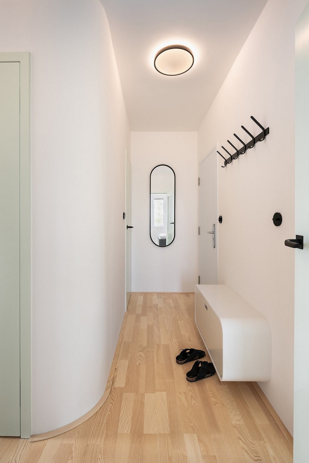 Modern entryway with white walls, wood flooring, circular mirror, and minimalist hooks.
