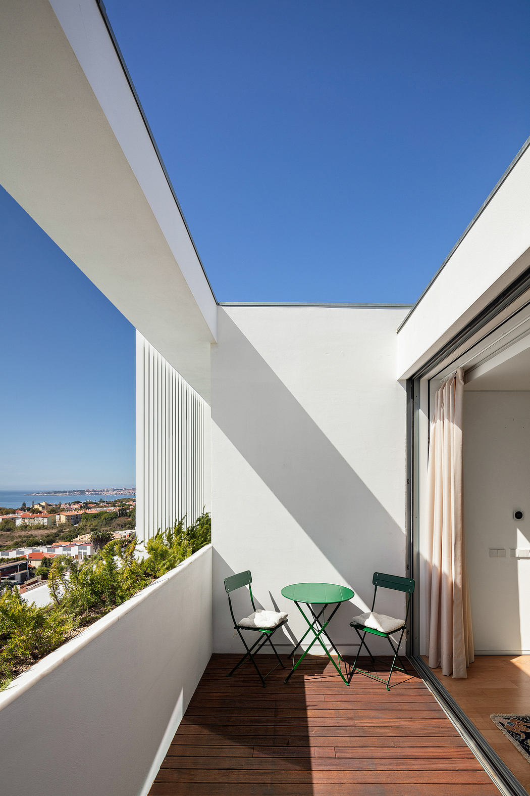 Minimalist balcony with wooden floor, green chairs, and panoramic city view through glass walls.