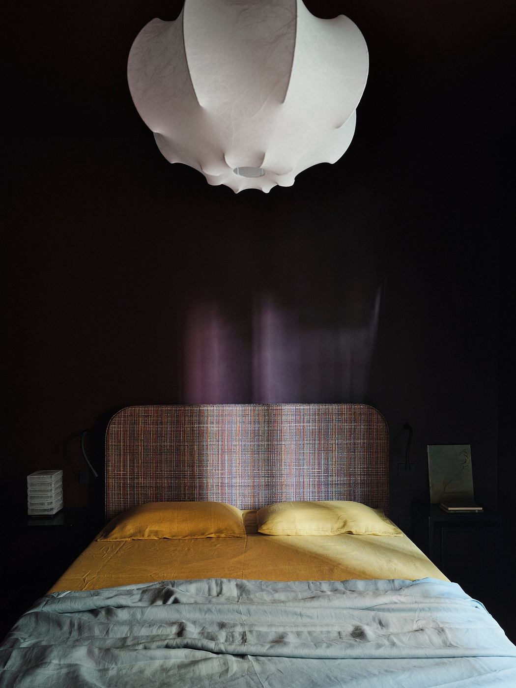 A dimly lit bedroom with a textured headboard, plush bedding, and an overhead light fixture.