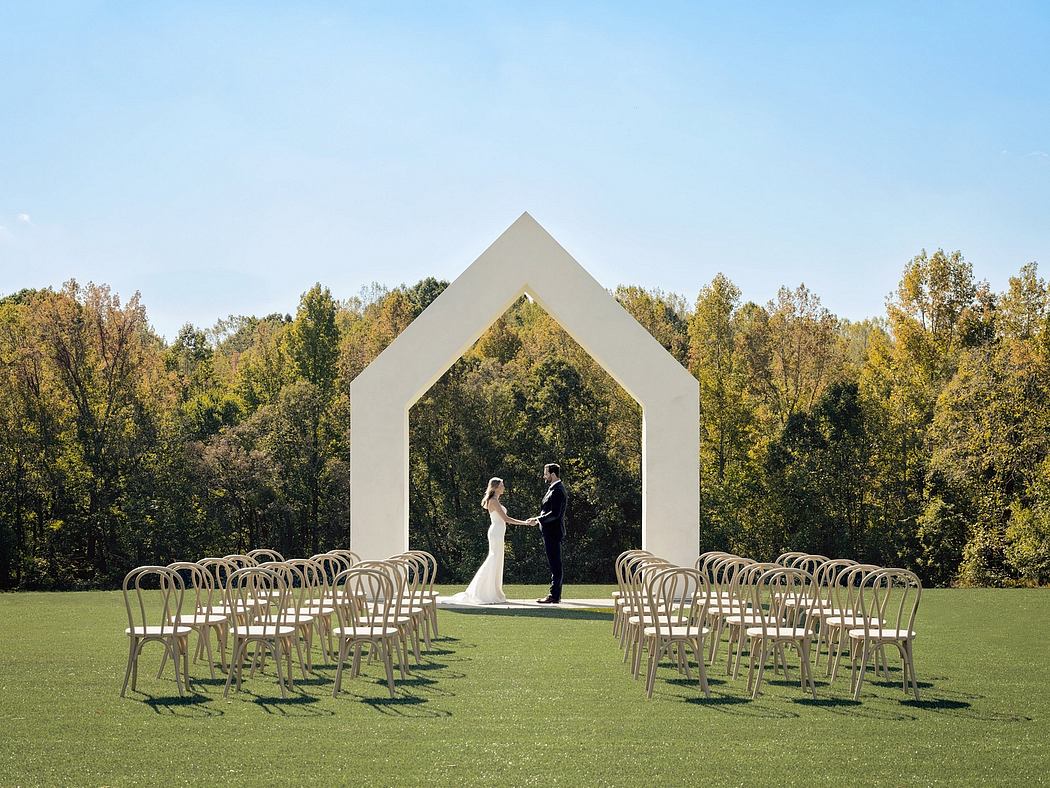 A white architectural archway frames a couple standing on a lush, grassy lawn surrounded by autumnal trees.