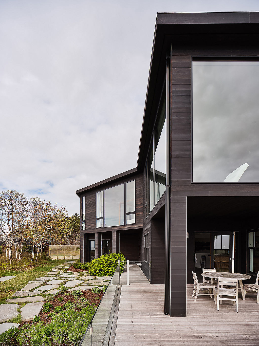 Sleek modern home with expansive windows, wood siding, and paved pathway leading to outdoor seating area.